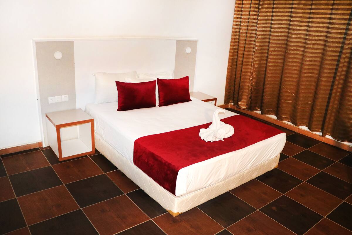 Una cama bien hecha con una manta y almohadas rojas, una mesita de noche y una ventana con cortinas.