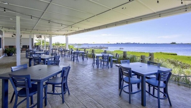 An outdoor dining area with tables and chairs, overlooking a body of water and a distant city skyline.