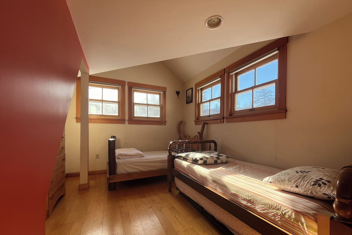 A small, cozy room with two wooden beds, a wooden dresser, and three windows providing natural light.