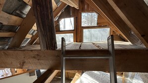 A loft bedroom with a wooden ladder, exposed beams, and large windows offering a view of the surrounding forest. - Streeter Mountain Farm (Rumney)