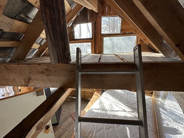 A loft bedroom with a wooden ladder, exposed beams, and large windows offering a view of the surrounding forest.