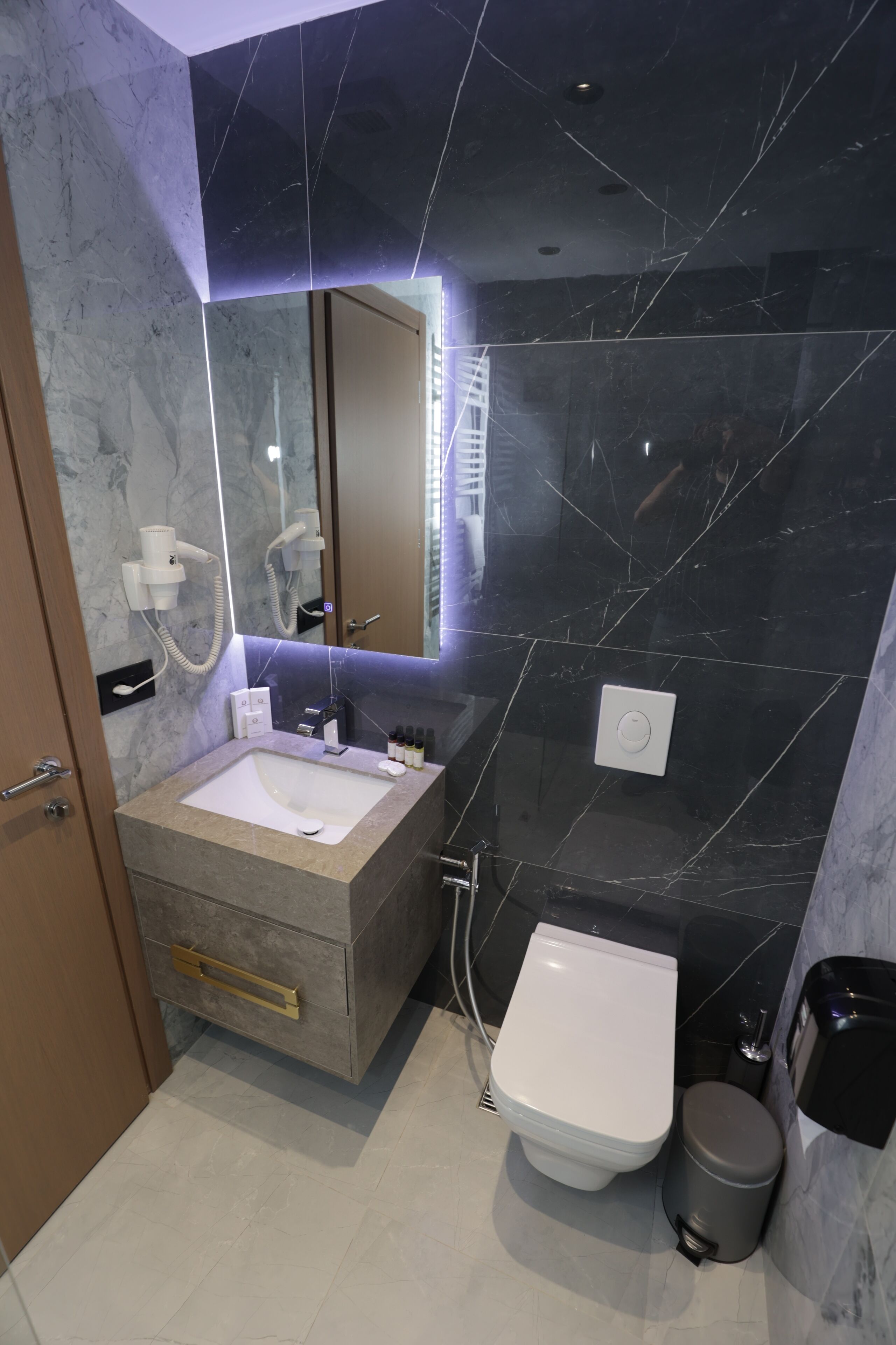 A modern bathroom with a marble vanity, a white sink, a toilet, and a trash bin.