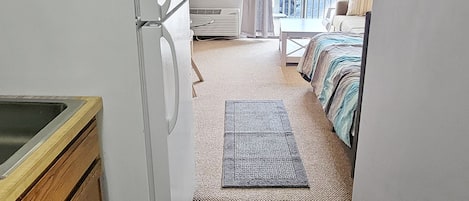 Cocina compacta con refrigerador, armarios de madera y un lavamanos pequeño. Se observa un comedor con mesa y sillas, que da a un balcón con vistas a edificios.