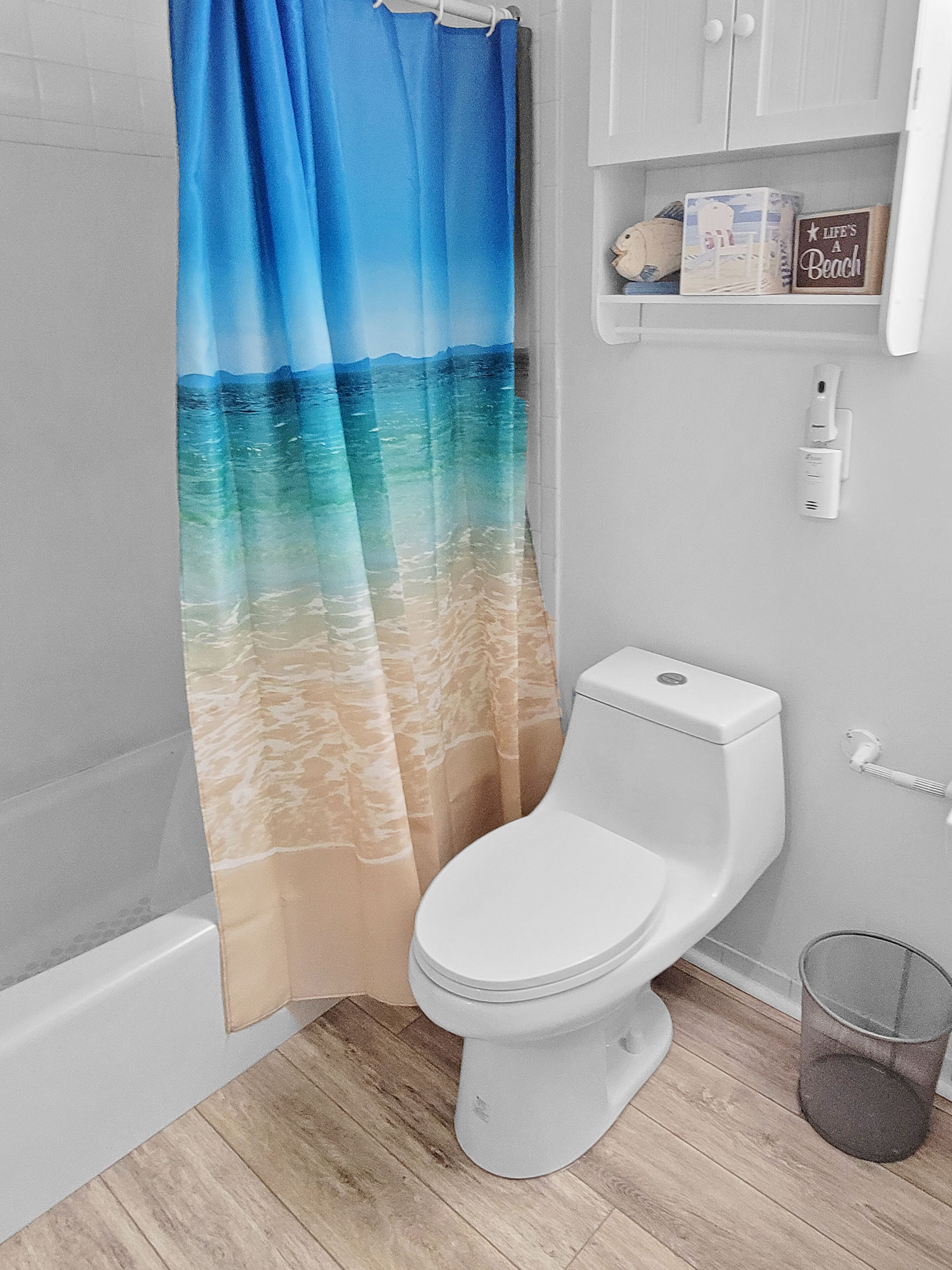 A bathroom with a toilet, a shower curtain depicting a beach scene, and a shelf with a 'Life's a Beach' sign.