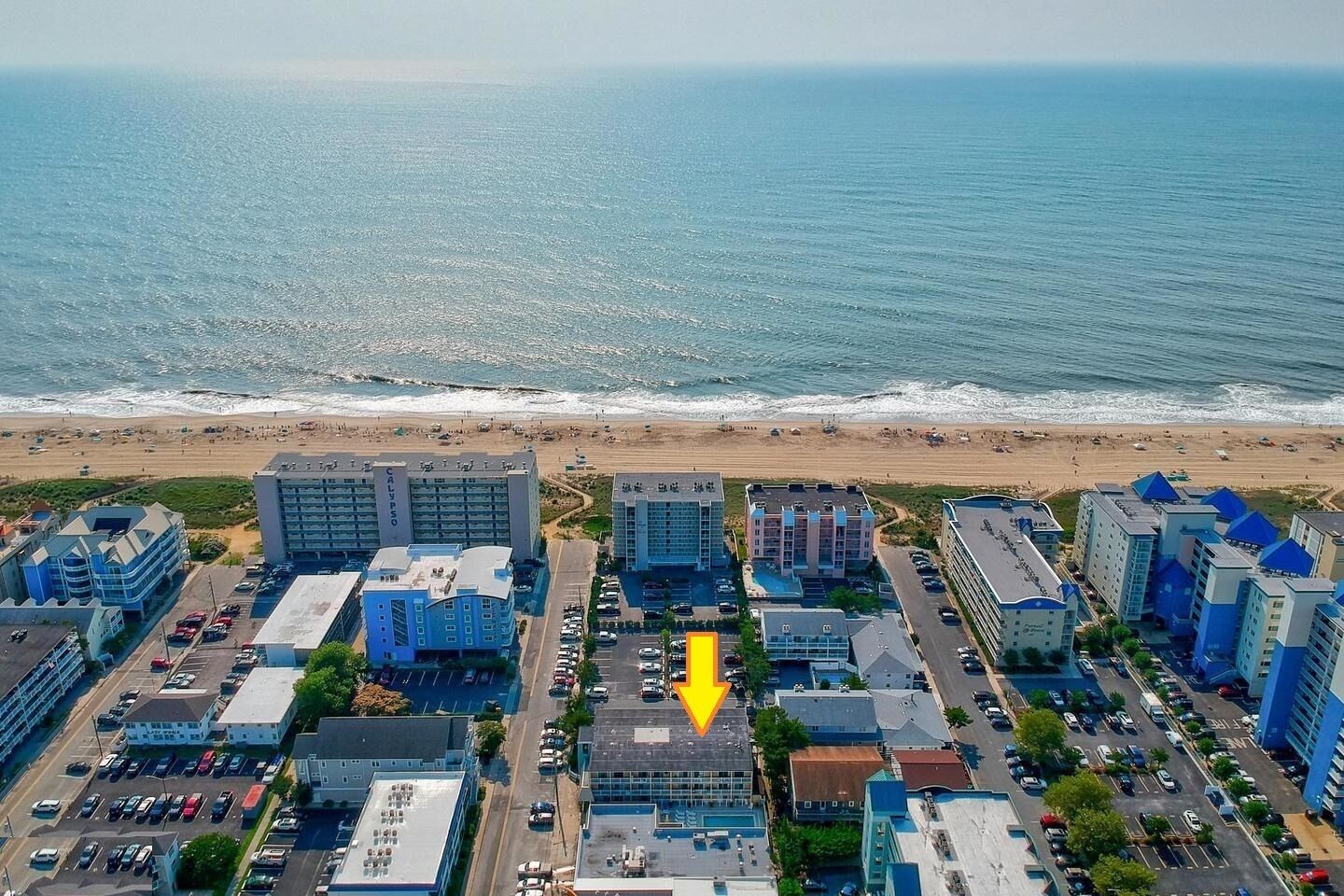 Vue aérienne d’un complexe hôtelier situé sur la côte, avec plusieurs bâtiments, un parking et une plage en arrière-plan.
