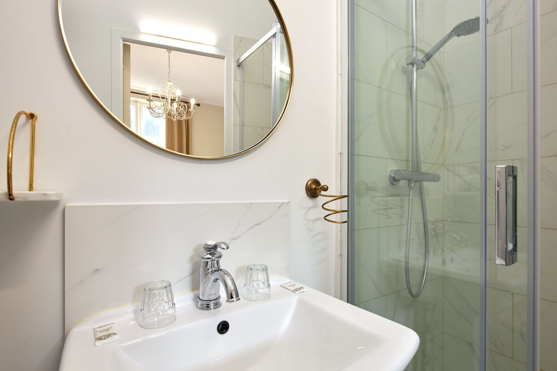 Une salle de bain avec un lavabo blanc, un miroir rond doté d’un cadre doré et une douche à parois de verre.