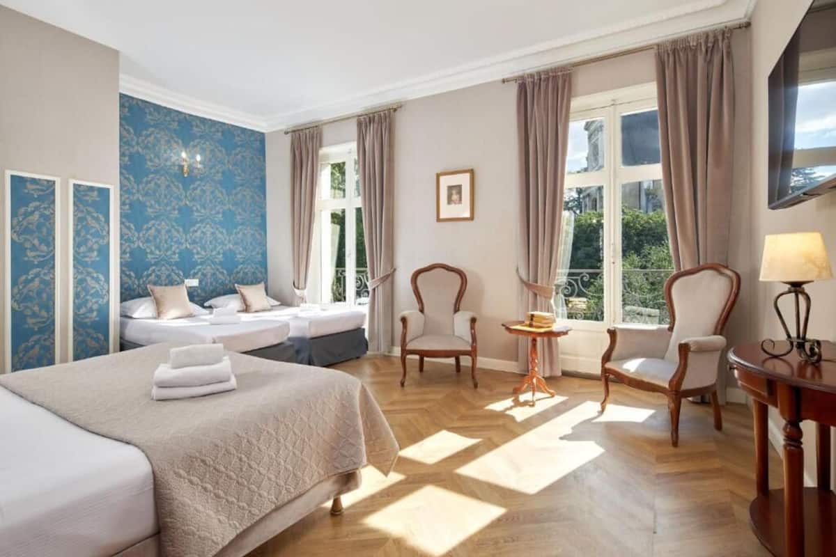 A hotel room with a large bed, two chairs, a small table, and a window with curtains.