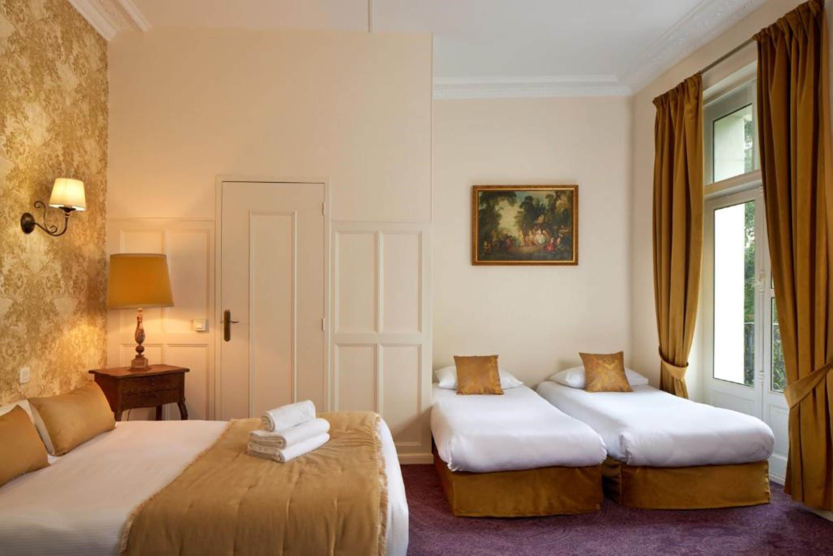 A hotel room with two single beds, a large window with curtains, a painting on the wall, and a lamp on a bedside table.