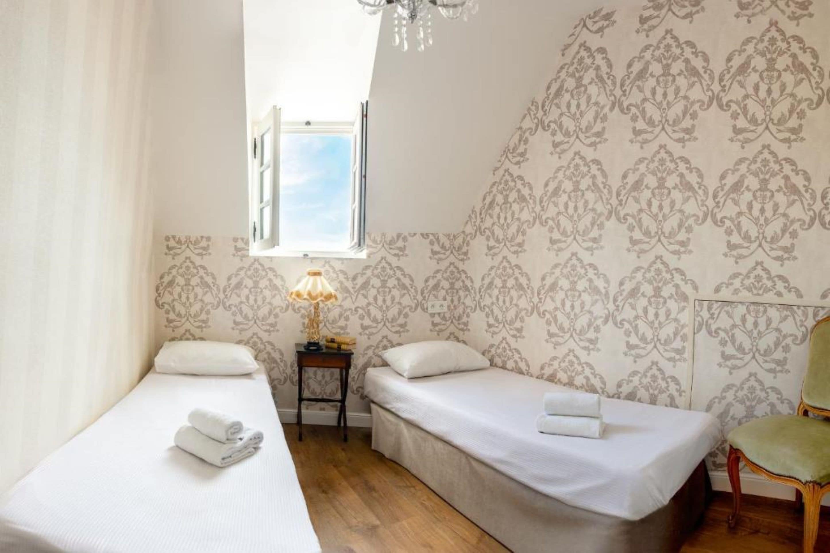 Una habitación pequeña y luminosa con dos camas individuales, una mesita de noche con lámpara, una silla y una ventana con cortinas.