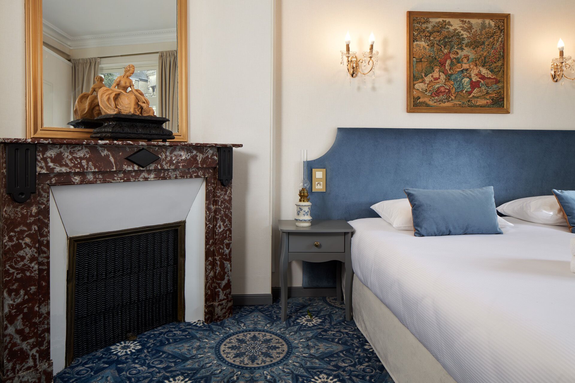 Une chambre avec une cheminée en marbre, un lit avec une literie bleue, une table de chevet avec une lampe et un tableau au mur.