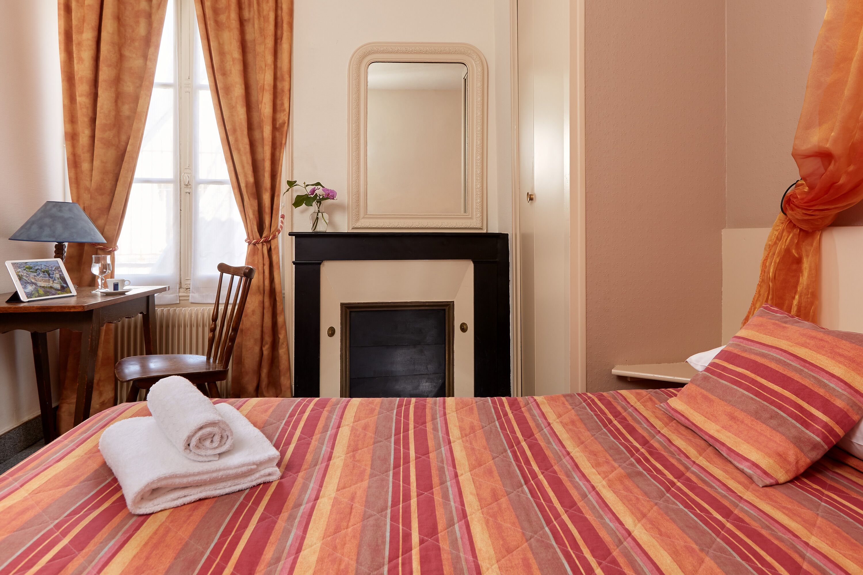 A hotel room with a bed, a chair, a desk with a lamp, a fireplace, and a window with curtains.