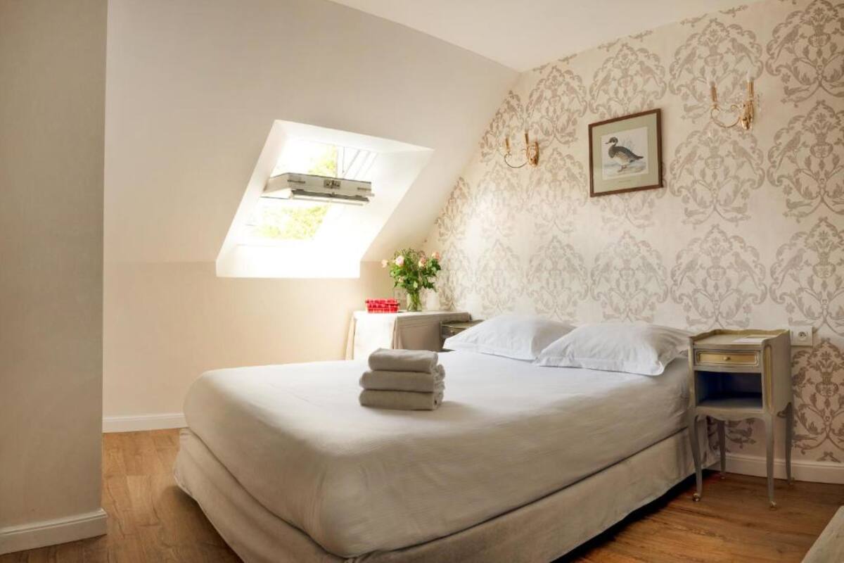 A bedroom with a large bed, a small table, a nightstand, a window with a plant, and a wall with a framed picture.