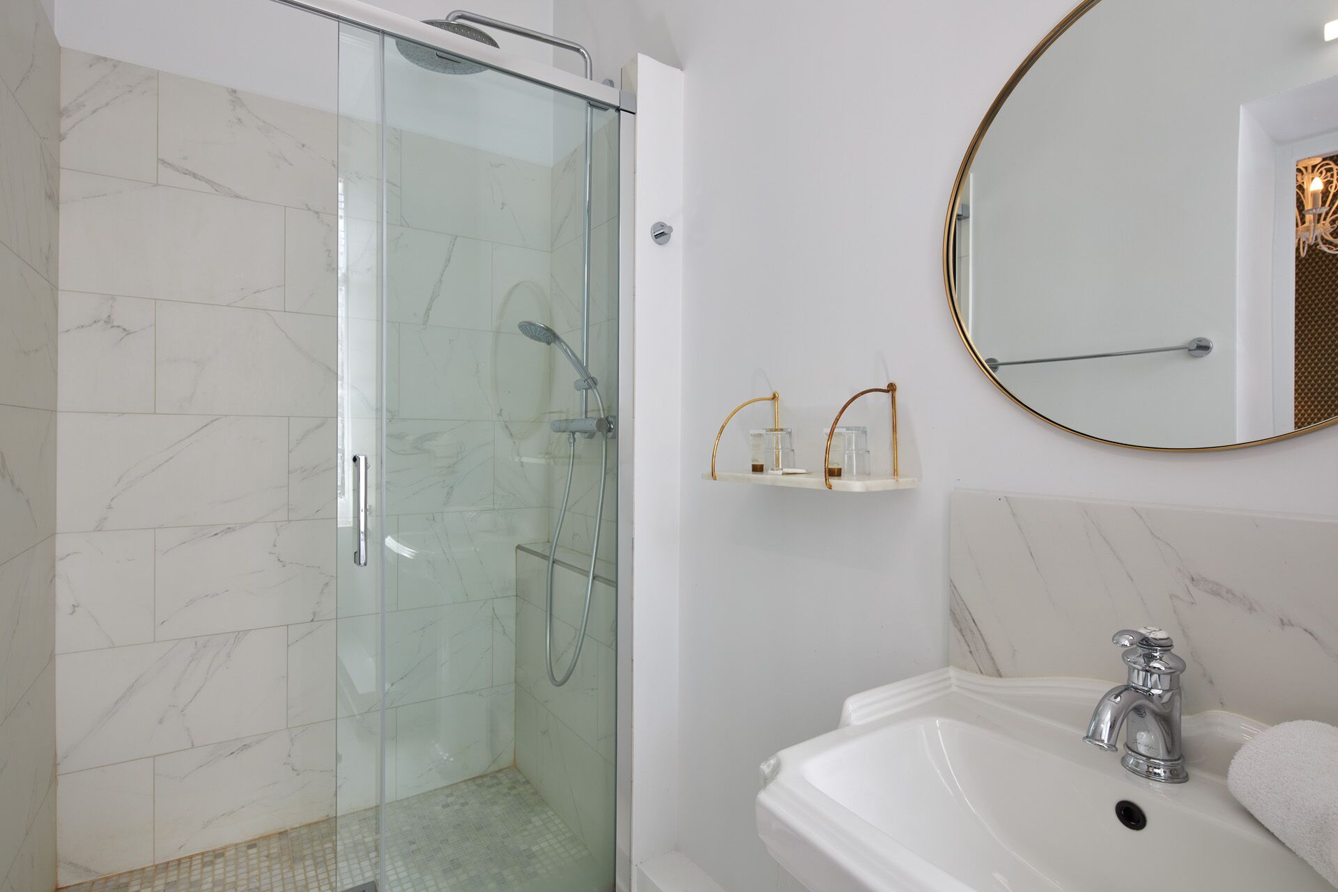 A bathroom with a glass-enclosed shower, a white sink with a single faucet, and a round mirror with a gold frame.