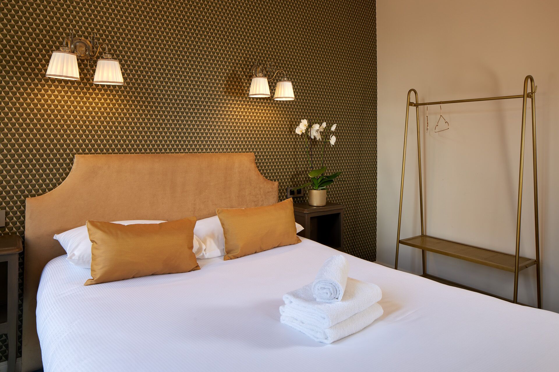 Een net opgemaakt bed met witte lakens en twee bruine kussens, een kleine bloeiende orchidee op een nachtkastje, en een spiegel met gouden lijst aan de muur.