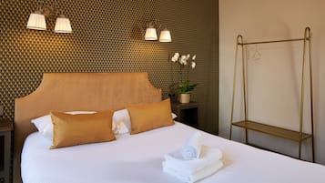 Een net opgemaakt bed met witte lakens en twee bruine kussens, een kleine bloeiende orchidee op een nachtkastje, en een spiegel met gouden lijst aan de muur.