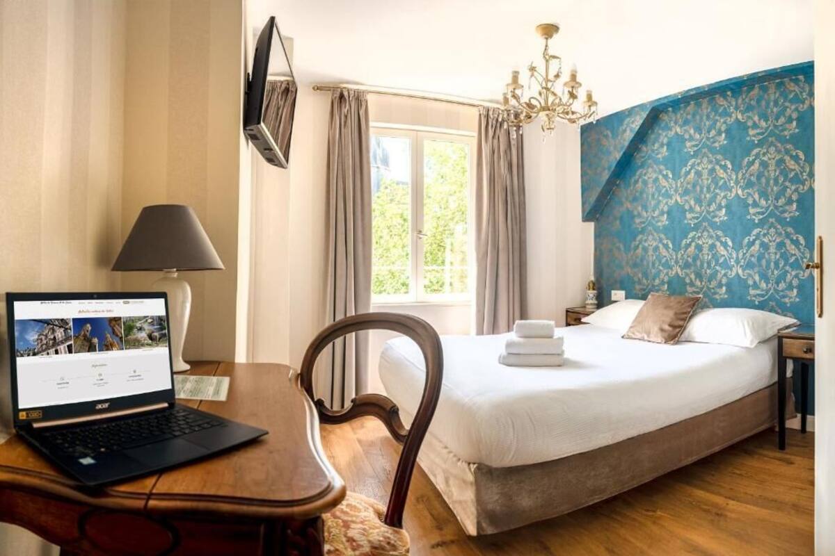 A hotel room with a bed, a desk with a laptop, a chair, a window with curtains, and a chandelier.