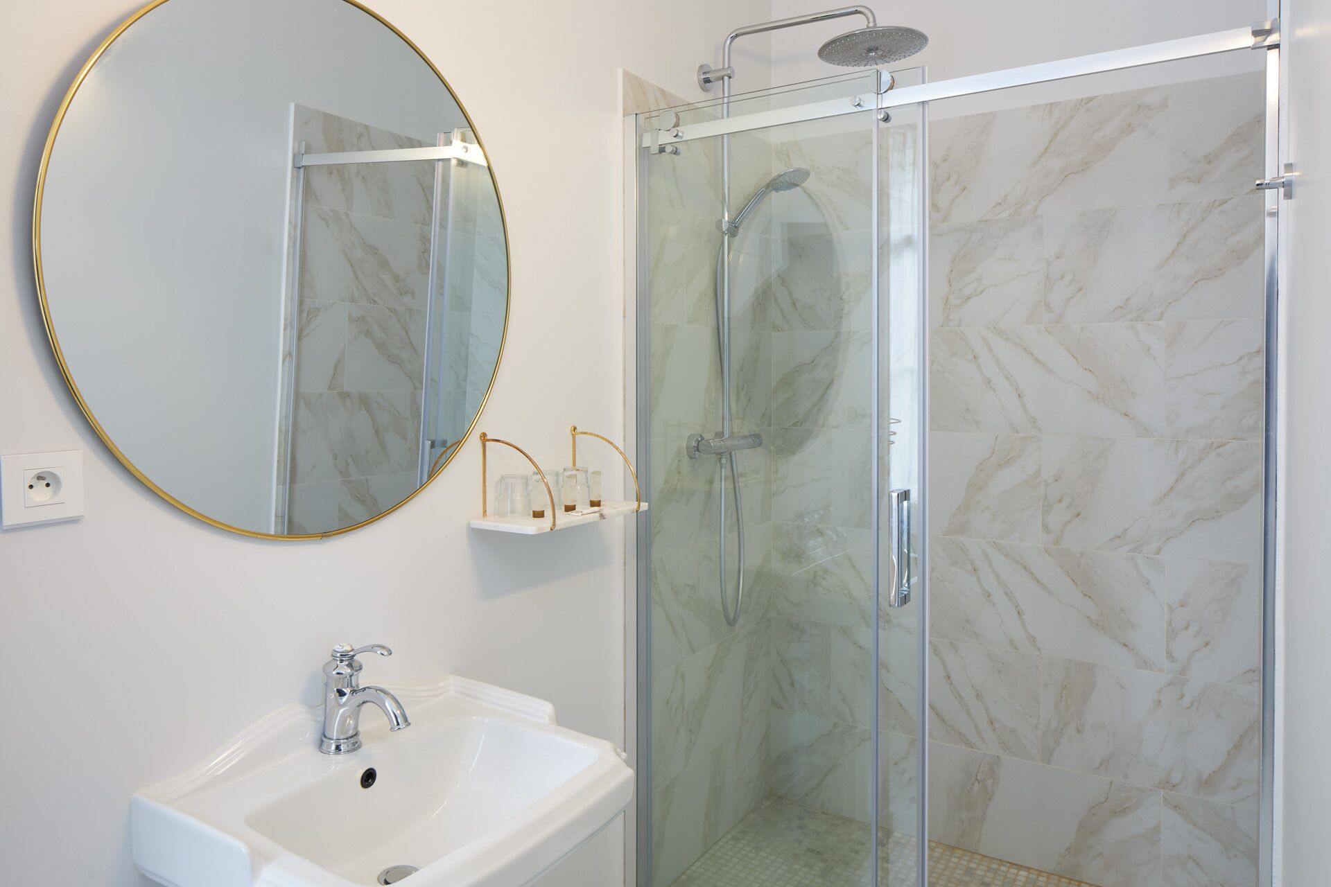 A bathroom with a glass-enclosed shower, a white sink, a round mirror with a gold frame, and a marble-patterned wall.