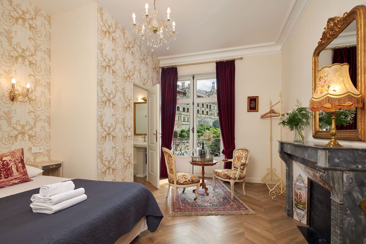 A hotel room with a bed, a sitting area with chairs, a fireplace, and a view of a historic building through the window.