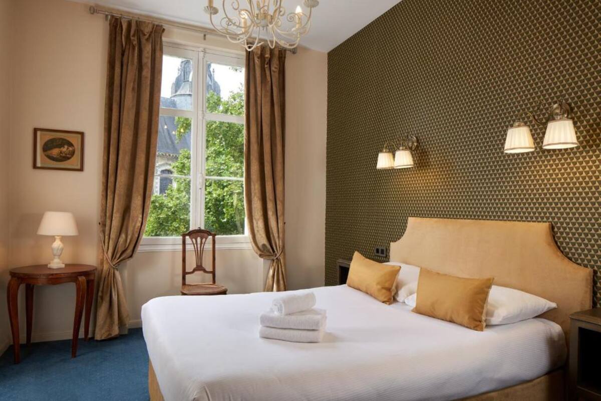 A hotel room with a large bed, a chandelier, a window with curtains, a small table with a lamp, and wall-mounted lights.