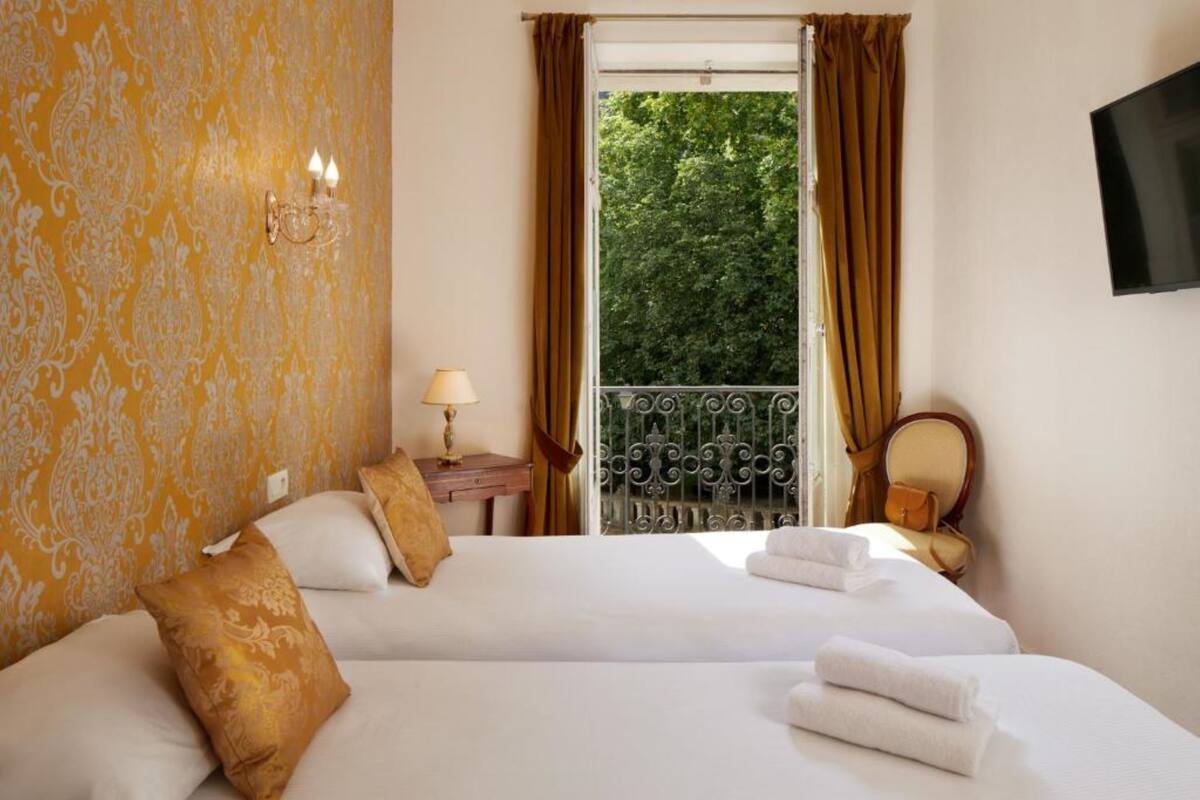 A hotel room with two beds, a balcony with a wrought iron railing, a television on the wall, and a patterned wallpaper.