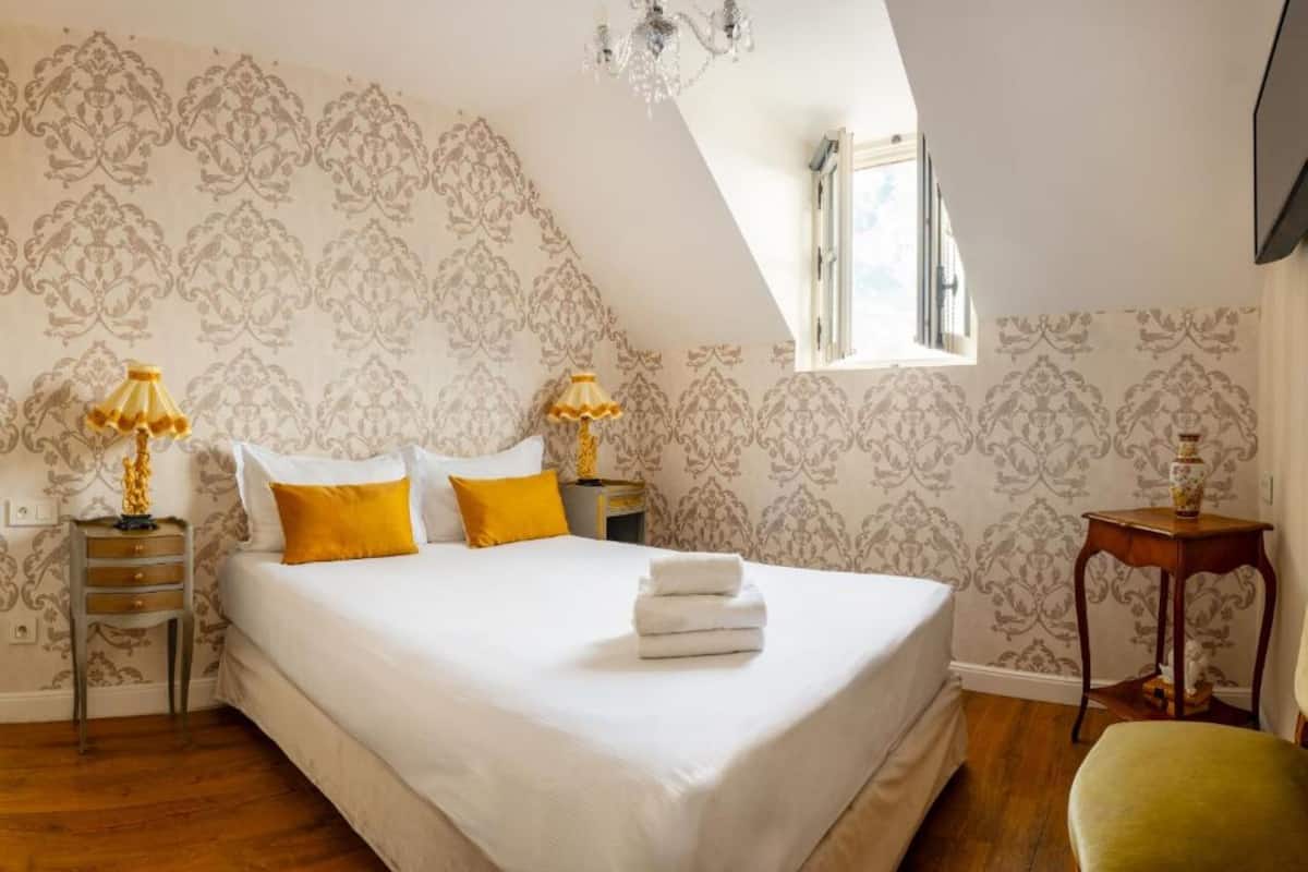 A bedroom with a large bed, two bedside tables with lamps, a window, and a small table with a vase.