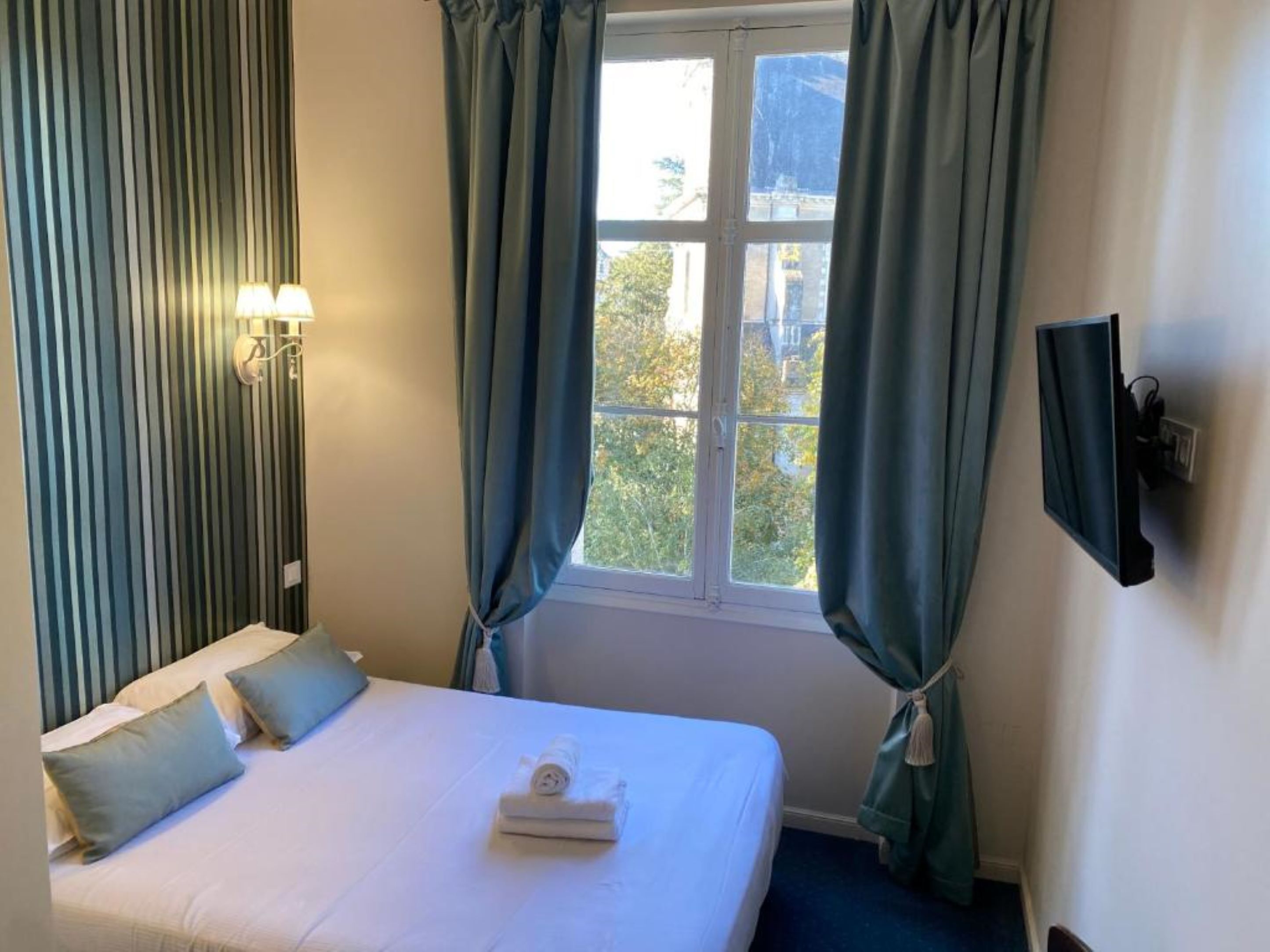 Une chambre d’hôtel avec un lit, une fenêtre avec des rideaux, un téléviseur fixé au mur et une lampe.