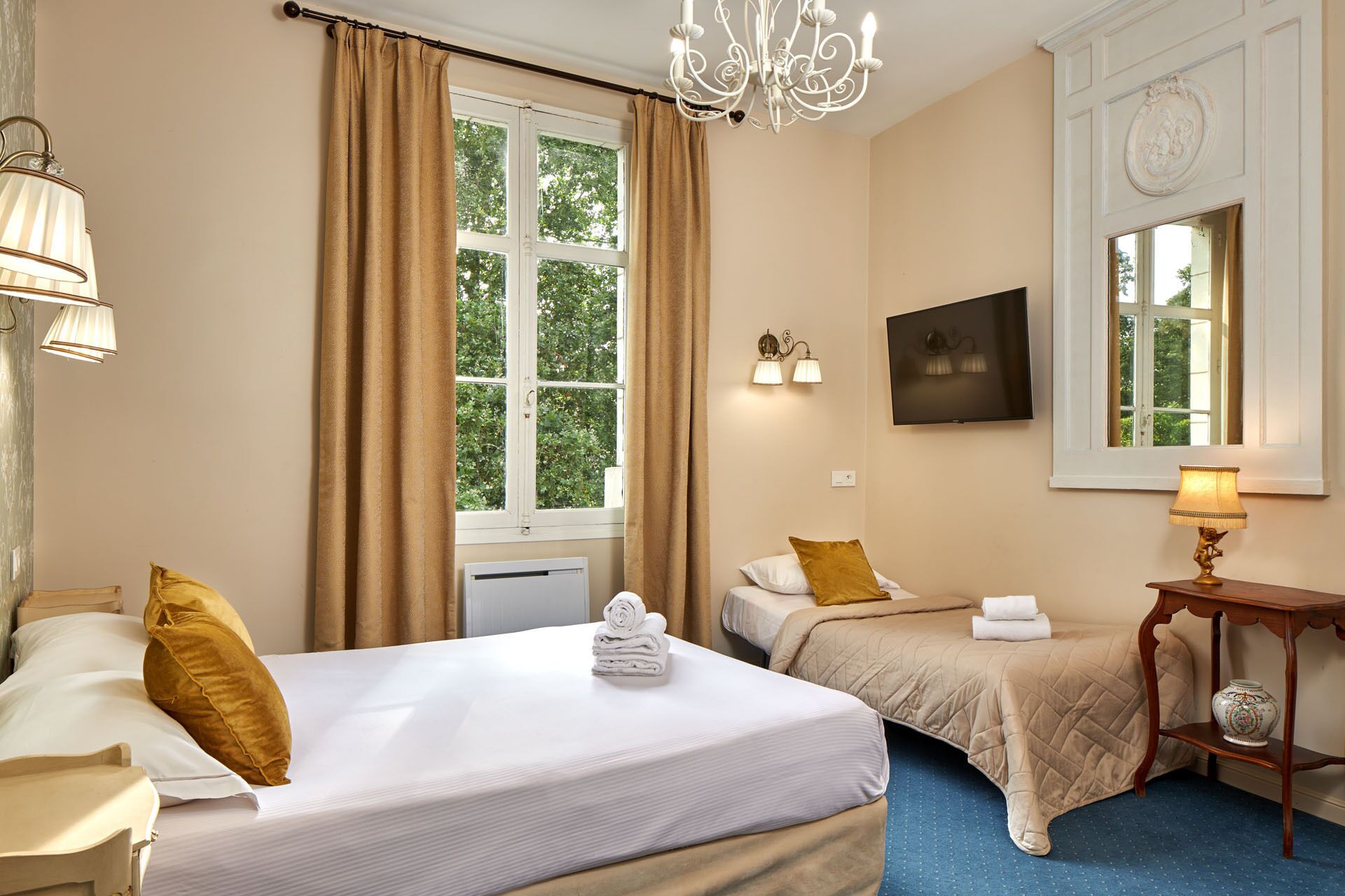 A hotel room with two beds, a TV, a chandelier, and a window with curtains.