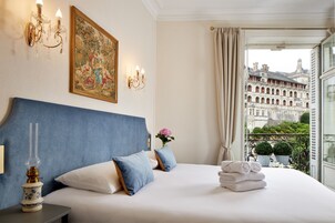 Une chambre d’hôtel avec un grand lit, un lustre, un tableau et une vue sur un bâtiment historique.
