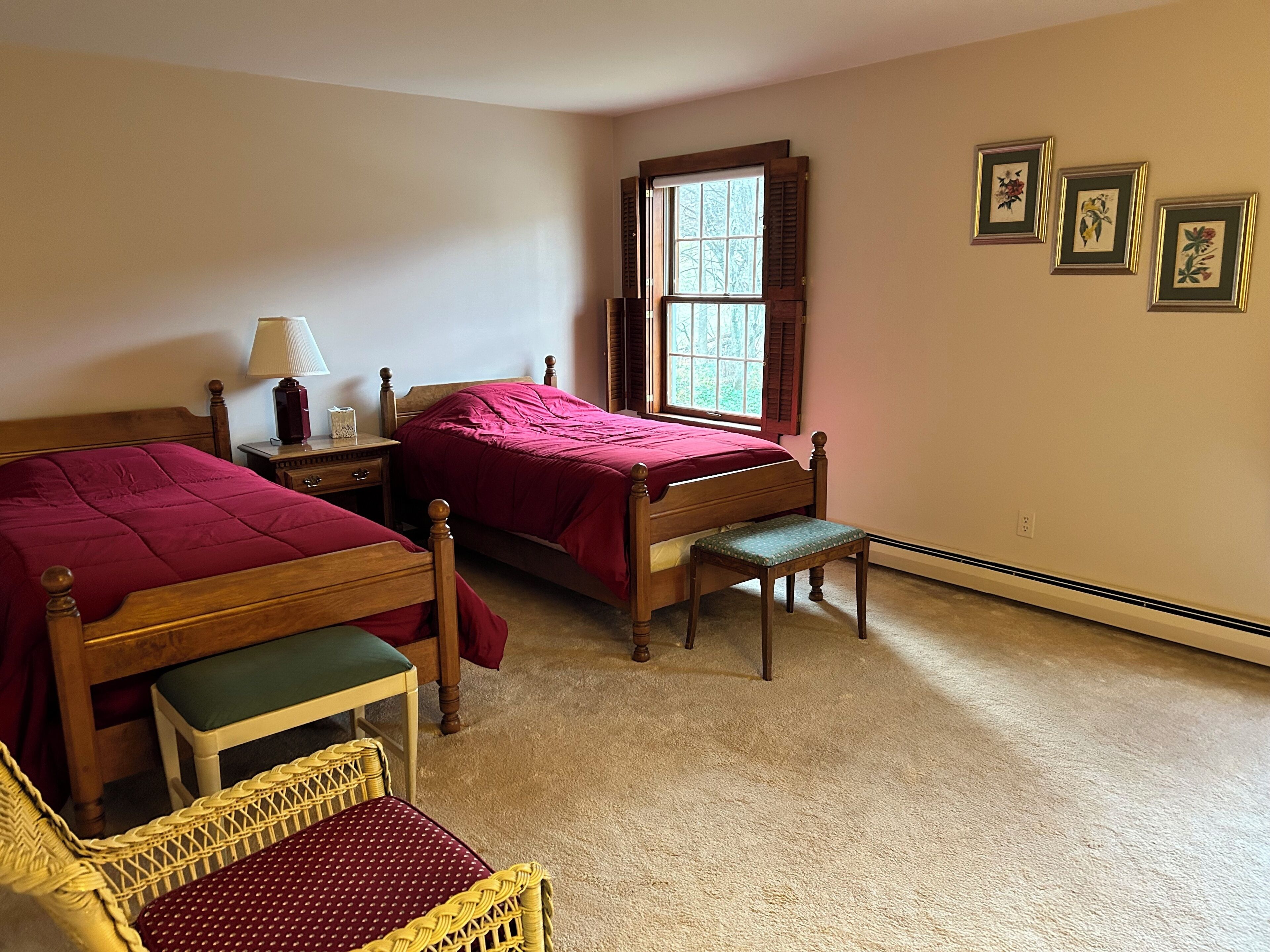 A bedroom with two wooden beds, a nightstand, a chair, a window with shutters, and three framed pictures on the wall.