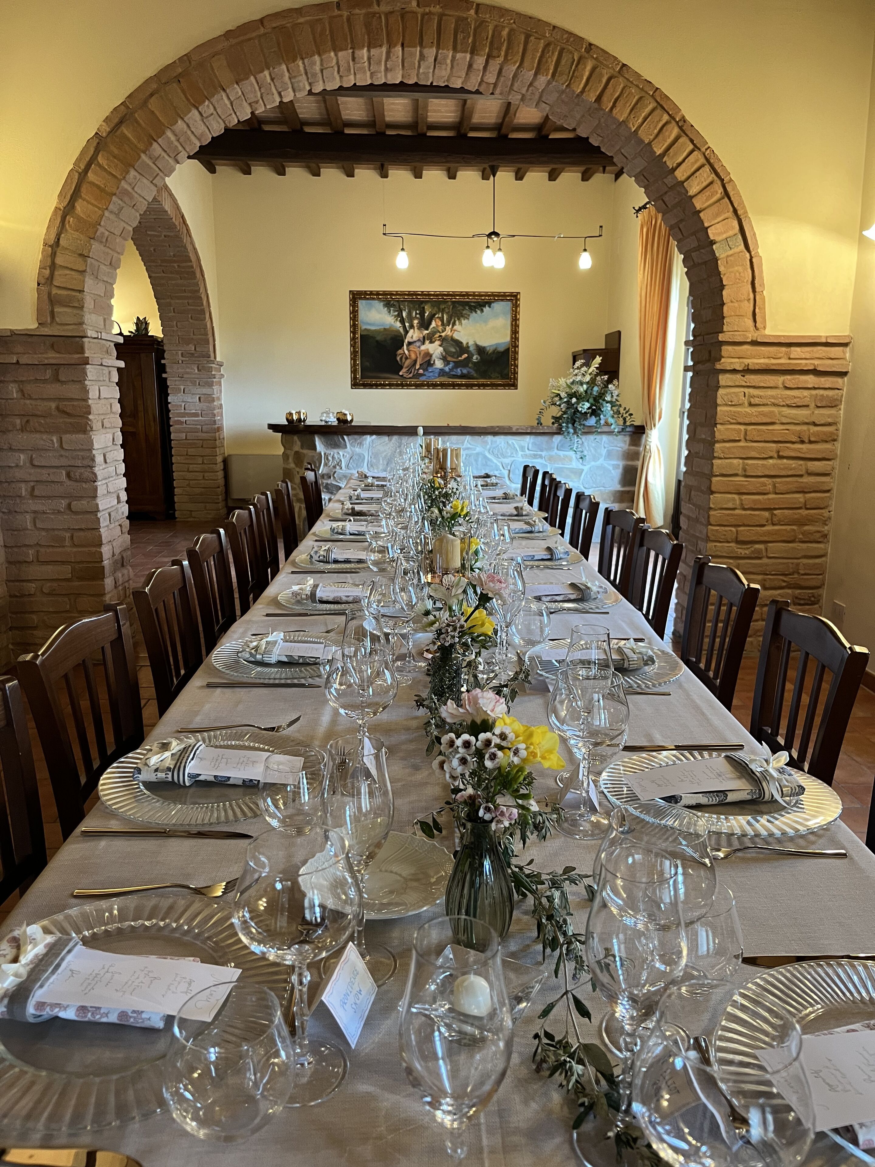A long dining table set for a formal meal, flanked by brick arches, with a painting above and a stone fireplace in the background.