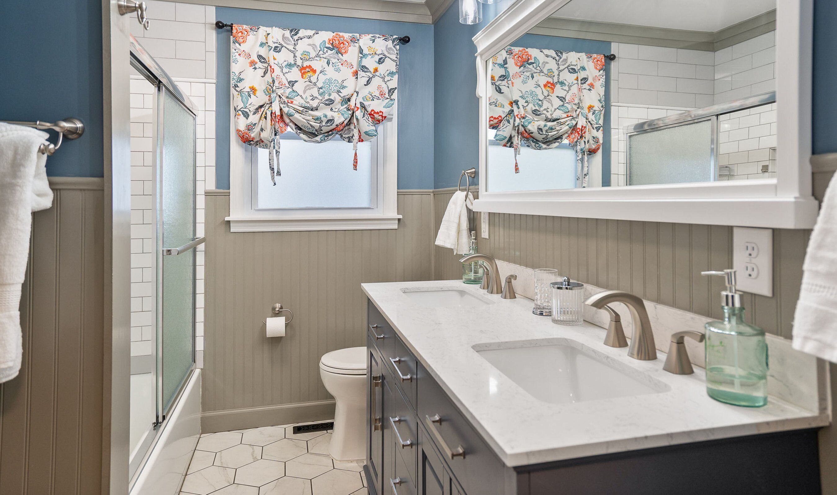 Un baño con lavamanos doble, inodoro, ducha y ventana con cortinas florales.