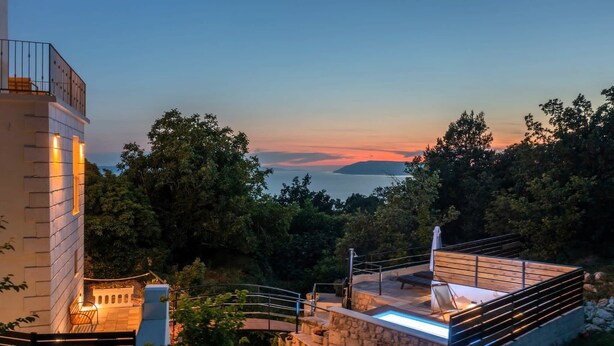 Un espace piscine extérieur moderne, agrémenté d’un mur en pierre et d’une terrasse en bois, entouré d’une végétation luxuriante et offrant une vue sur une étendue d’eau.