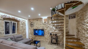 Un salon avec un mur en pierre, un canapé, un téléviseur, un escalier en bois et une petite table sur laquelle est posée une plante.