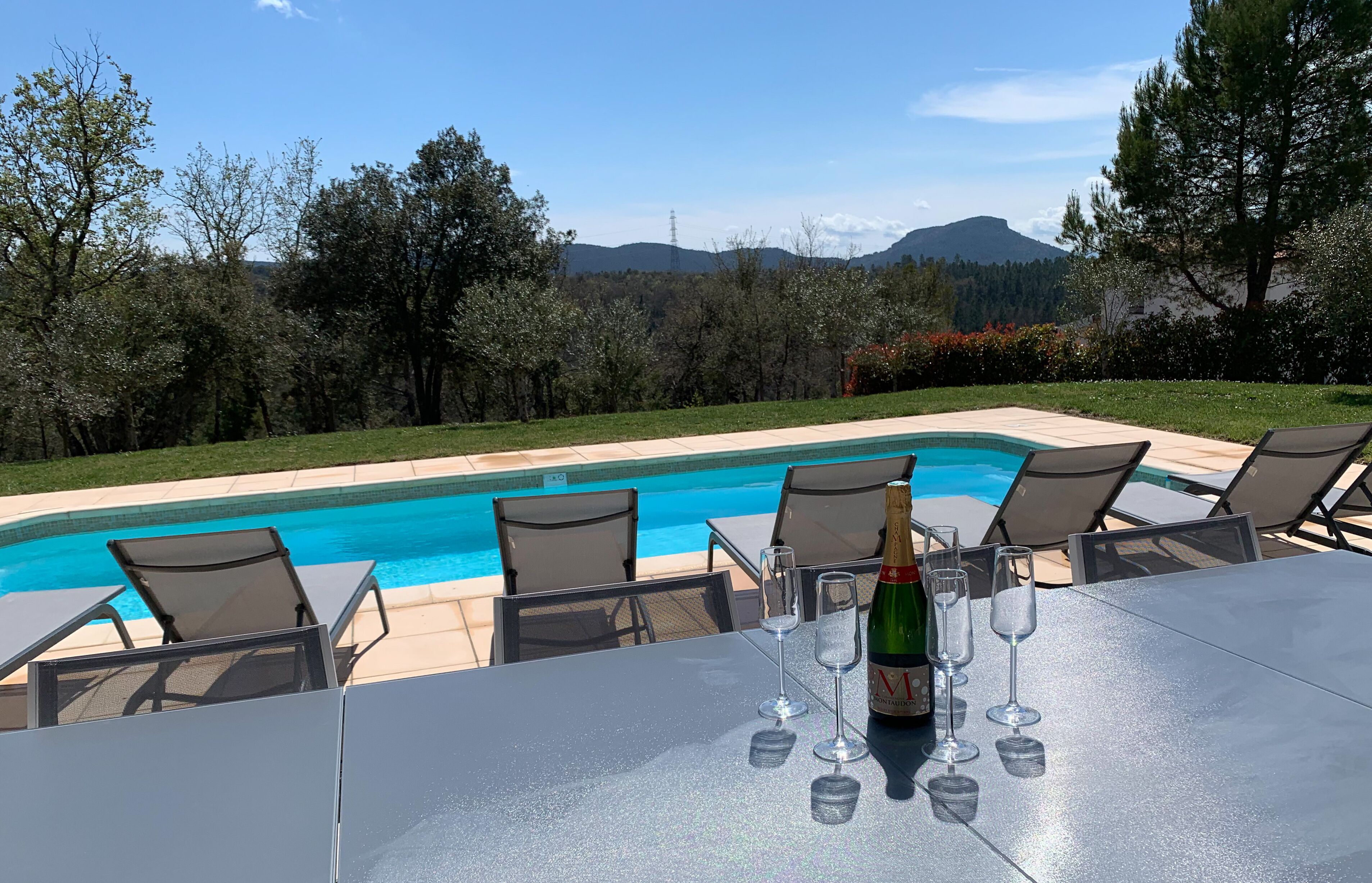 Une table placée au bord de la piscine, avec des chaises et des coupes à champagne, offrant une vue sur la piscine et sur les montagnes.