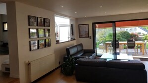En moderne stue med en sort lædersofa, et spiseområde med bord og stole, og et stort vindue der giver udsigt til naturen.