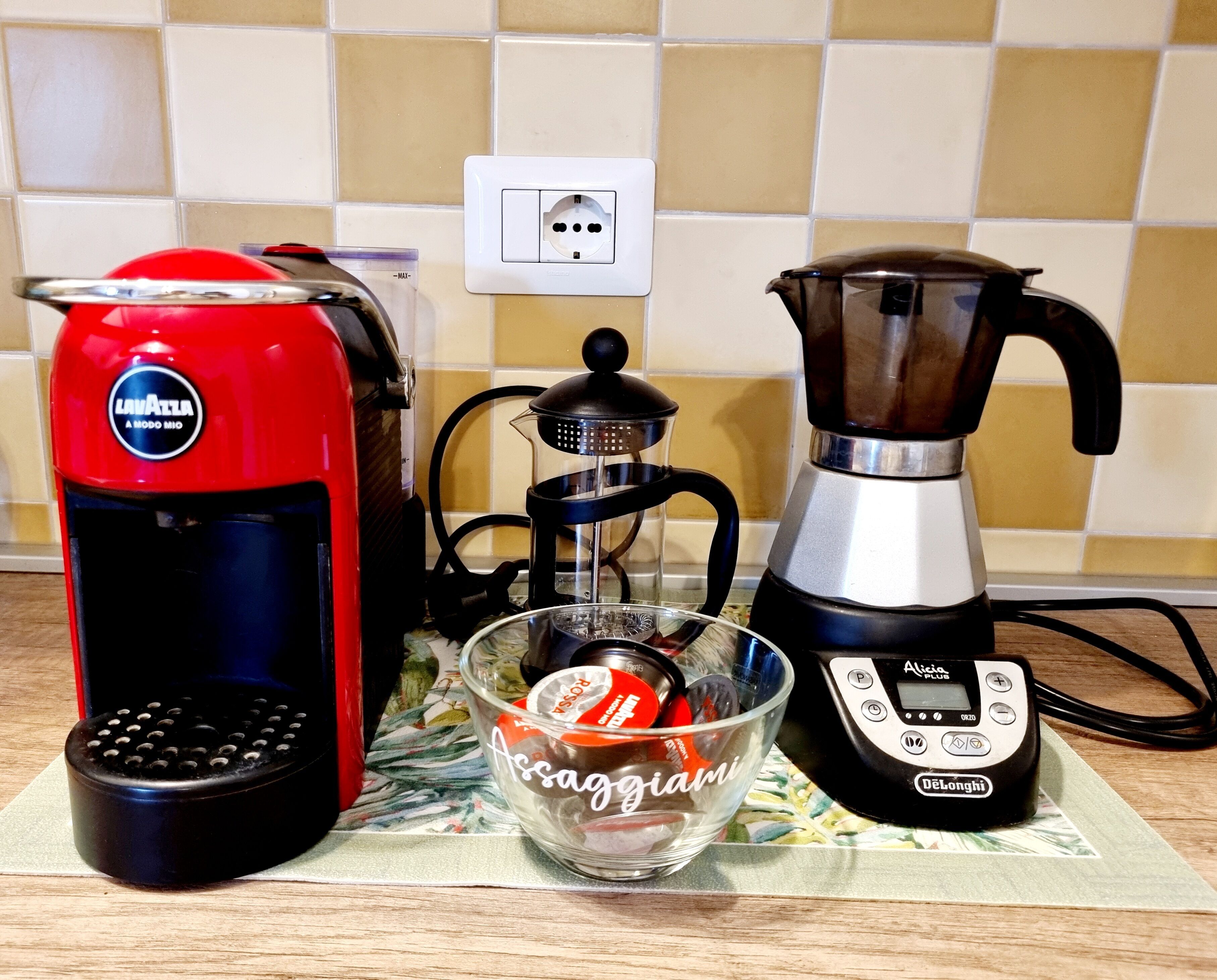 A kitchen counter with a coffee maker, a coffee pot, a coffee grinder, and a glass container with coffee pods.