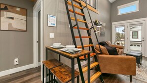 A modern living room with a wooden table set for dining, a brown leather sofa, a black ladder, and a window with a view of the outside. - Luxury Living – Walk to Poudre Trail and Old Town! (Fort Collins)
