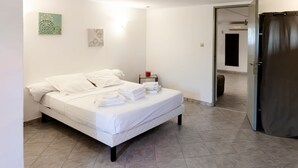 Una cama bien hecha con sábanas y toallas blancas, una mesita de noche con una taza roja y una puerta que da a otra habitación.