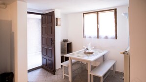 Una habitación pequeña con una mesa blanca y bancos, una puerta de madera, un radiador y una ventana con cortinas.