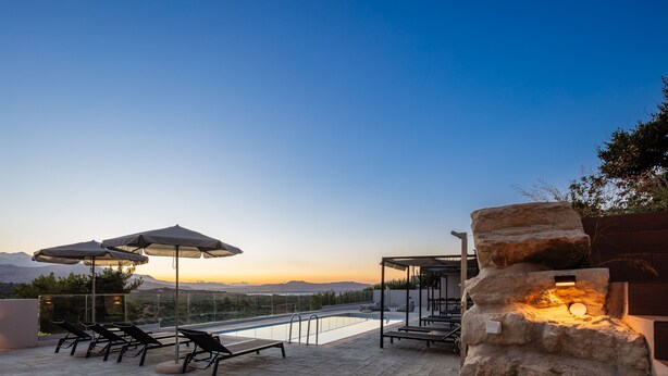 An outdoor swimming pool area with lounge chairs and umbrellas, surrounded by a stone wall and overlooking a mountainous landscape. - Suites Olive Tree Farm Episkopi Village (Rethymno)