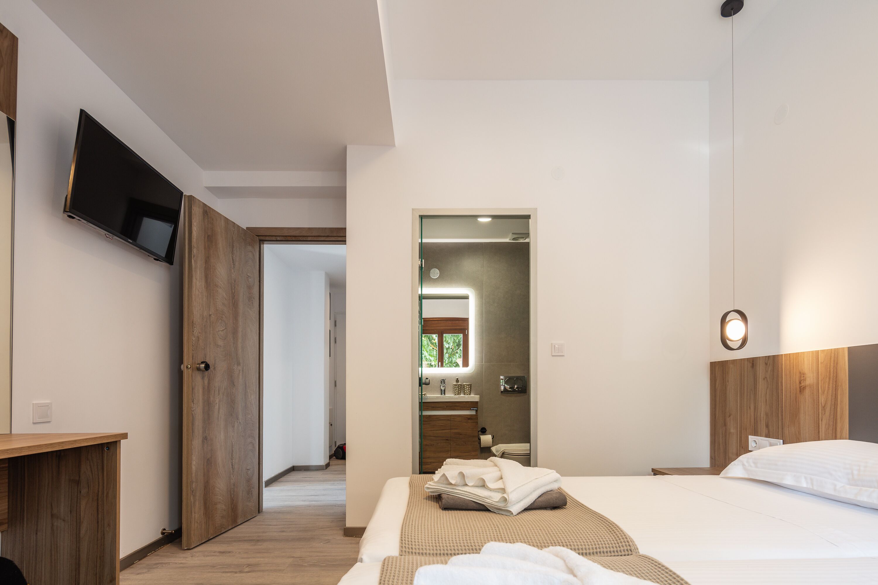 A modern hotel room with a wooden desk, a flat-screen TV, a large bed with white linens, and a bathroom visible through an open door.