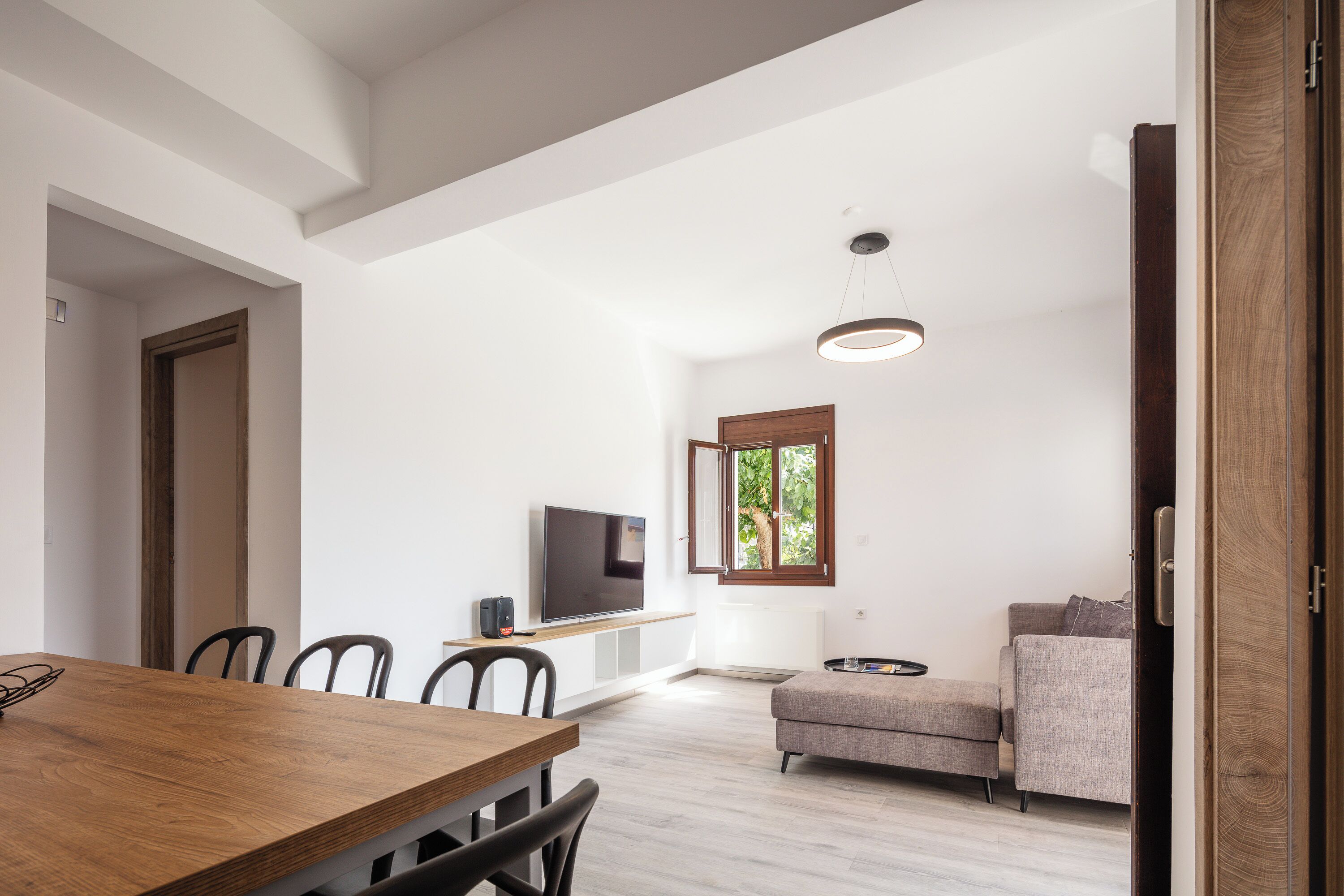 Een moderne woonkamer met een houten eettafel, een bank, een televisie en een raam met uitzicht op bomen.