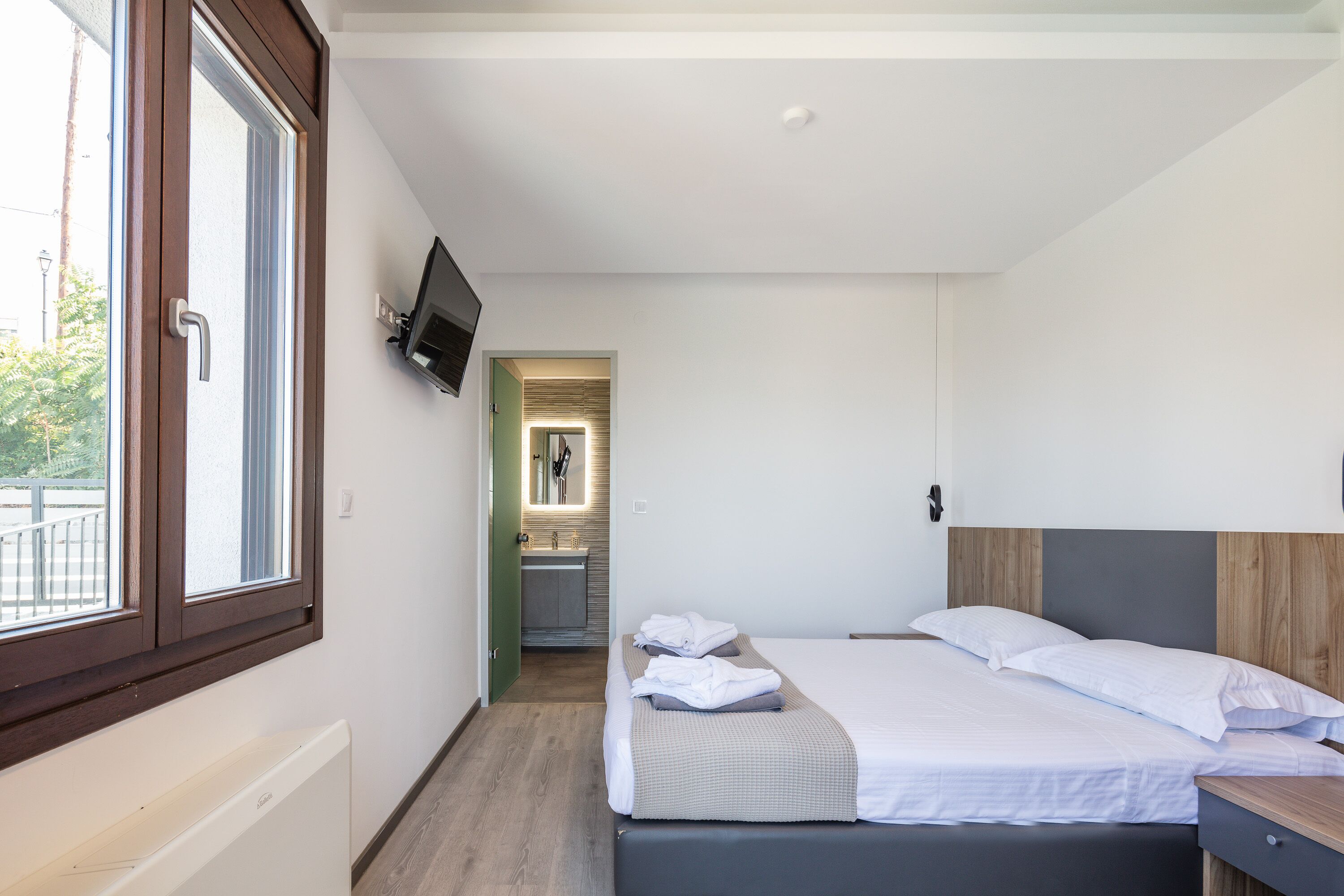 Quarto de hotel moderno com uma cama grande, uma mesa de cabeceira, uma TV na parede e vista para o banheiro através de uma porta aberta.