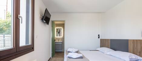 Quarto de hotel moderno com uma cama grande, uma mesa de cabeceira, uma TV na parede e vista para o banheiro através de uma porta aberta.