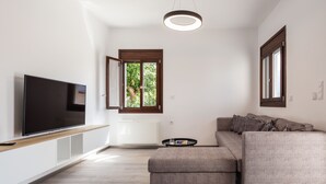 A modern living room with a large flat-screen TV, a grey sectional sofa, and a window with an open shutter. - Suites Olive Tree Farm Episkopi Village (Rethymno)