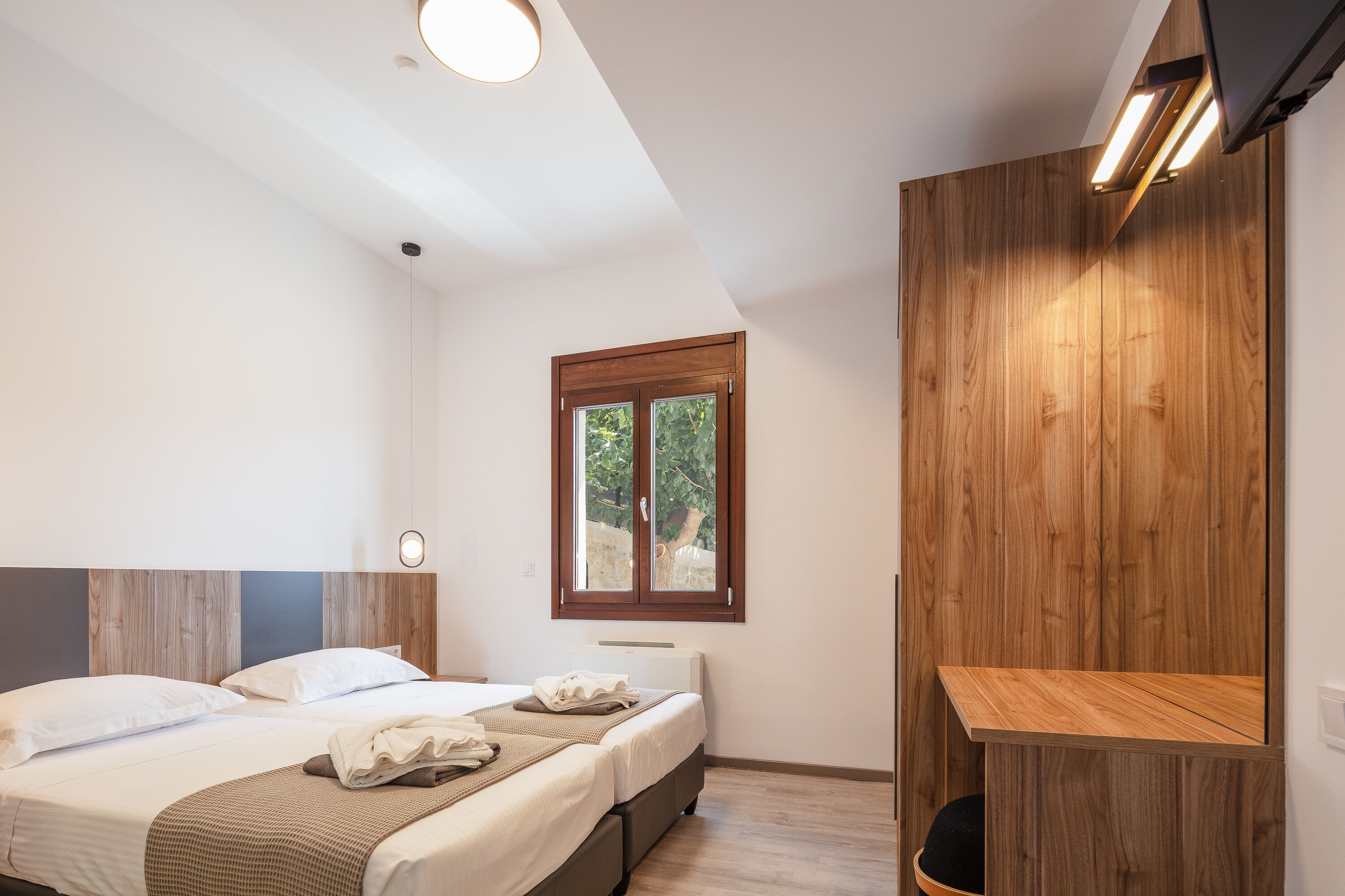 A modern bedroom with two beds, a wooden accent wall, a window with a view, and a built-in TV shelf.
