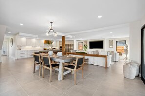 A modern kitchen with white cabinets, a dining area with a wooden table and chairs, and a living area with a sofa and a TV. - Gorgeous Waterfront Luxury Beach House Sleeps 22, Pool, Kayaks (Panama City Beach)