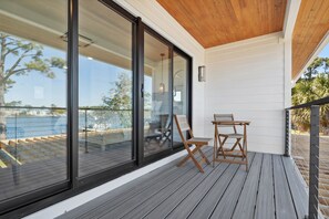 A covered outdoor deck with a wooden floor, two wooden chairs, and a glass door leading to an indoor area. - Gorgeous Waterfront Luxury Beach House Sleeps 22, Pool, Kayaks (Panama City Beach)