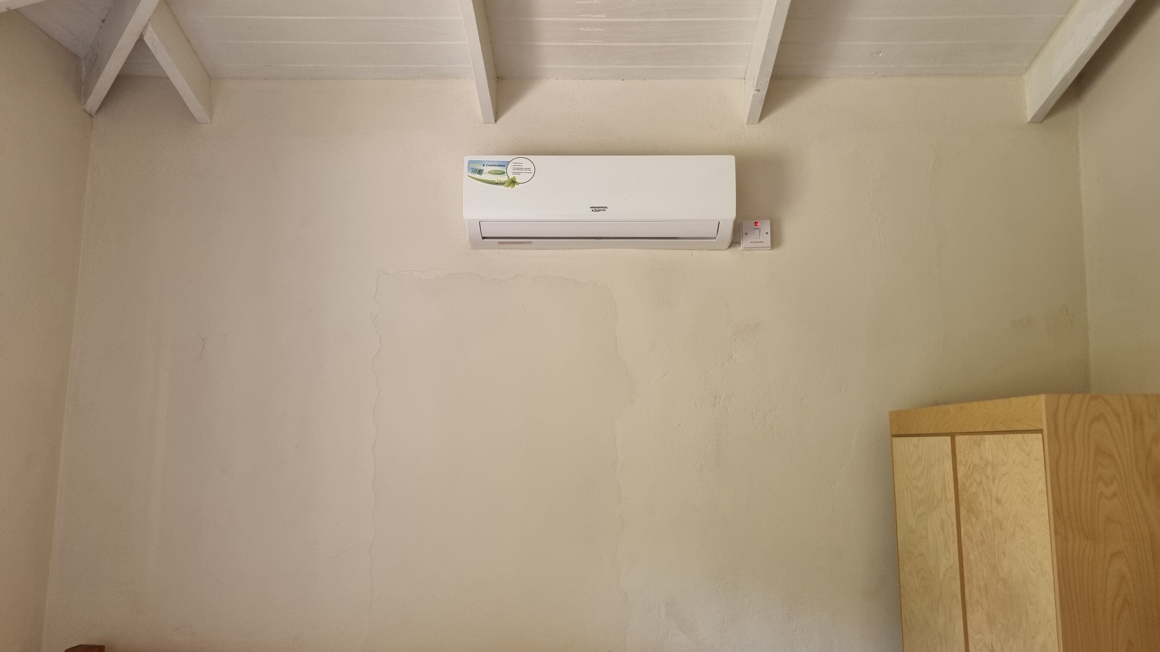 A room with a white ceiling, a white air conditioning unit mounted on the wall, and a wooden cabinet to the right.