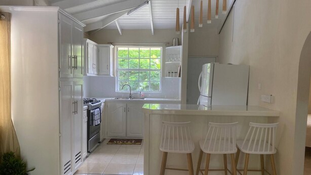 A kitchen with white cabinets, a stove, a sink, and a refrigerator. There are three white bar stools at the counter. - 2 bedroom house  (Vieux-fort)